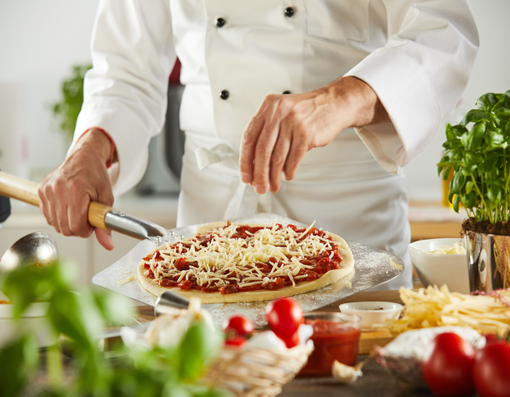 Chef preparing a homemade pizza by sprinkling shredded cheese over tomato sauce in a professional kitchen with fresh ingredients